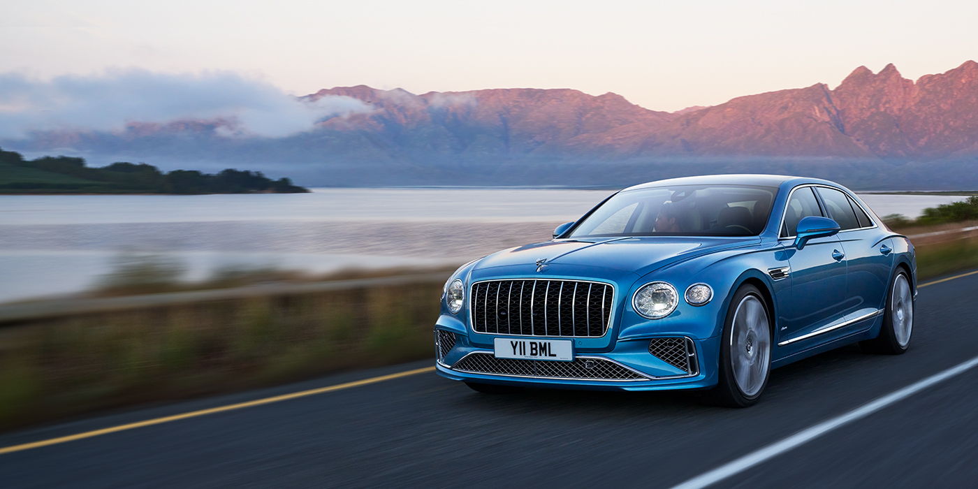 Bach Premium Cars GmbH New Bentley Flying Spur Azure sedan in Light windsor blue paint by Mulliner, with 22 inch Azure wheels and chrome straight vaned grille driving on a mountain road