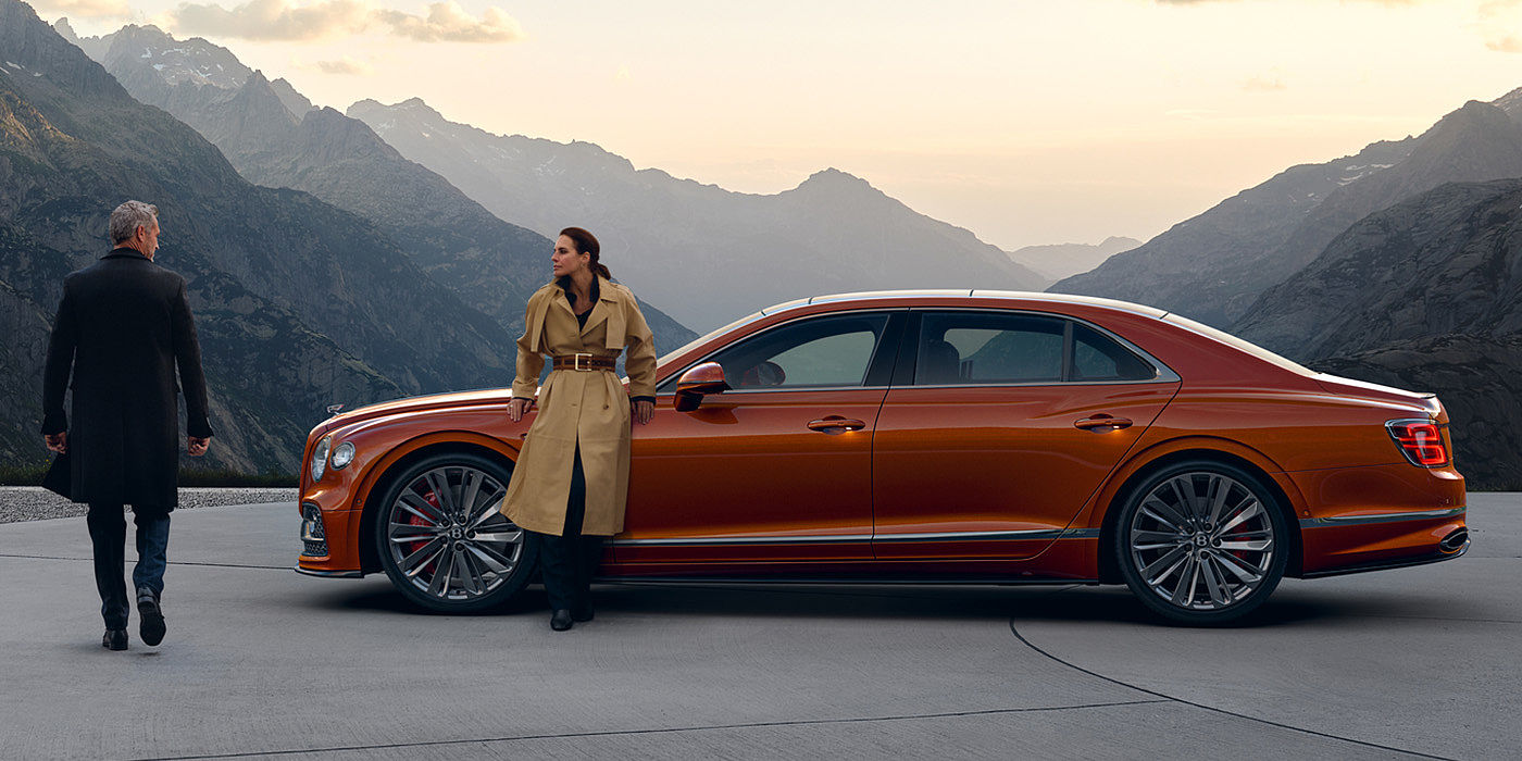Bach Premium Cars GmbH Bentley Flying Spur Speed parked in Orange Flame coloured exterior parked, with mountainous background and two people in view.