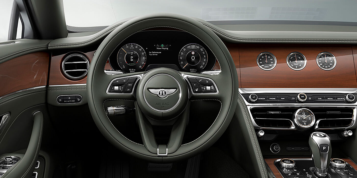 Bach Premium Cars GmbH New Bentley Flying Spur sedan front cabin in Cumbrian Green hide and Crown Cut walnut veneer showing steering wheel and Rotating Display with three clocks on face
