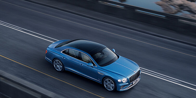 Bach Premium Cars GmbH New Bentley Flying Spur Azure sedan in Light windsor blue paint by Mulliner, with 22 inch Azure wheels seen from overhead driving dynamically on a country road